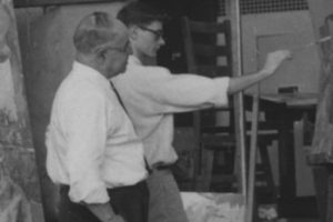 An older Jewish man in a shirt and tie observes a younger man with glasses painting on a vertical surface indoors; chairs behind them. *AI generated alt text